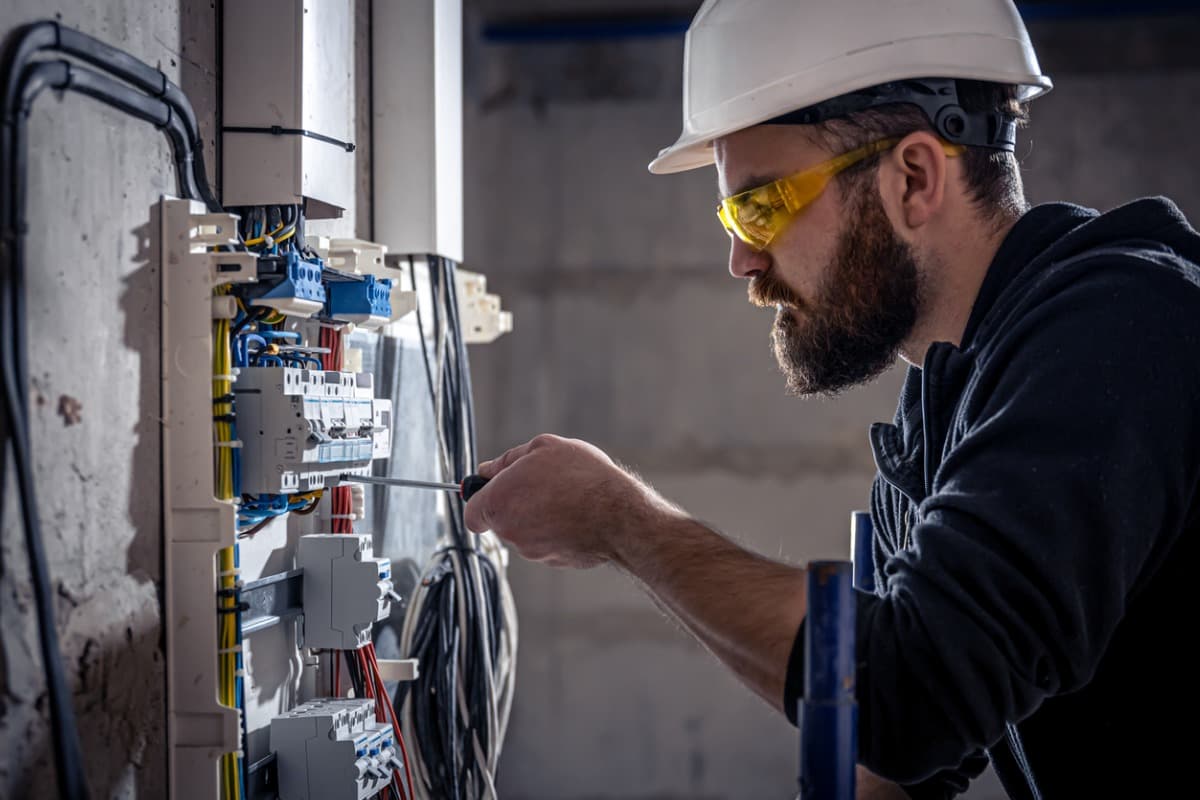 Électricien barbu avec casque blanc et lunettes jaunes réparant un tableau électrique avec un tournevis.